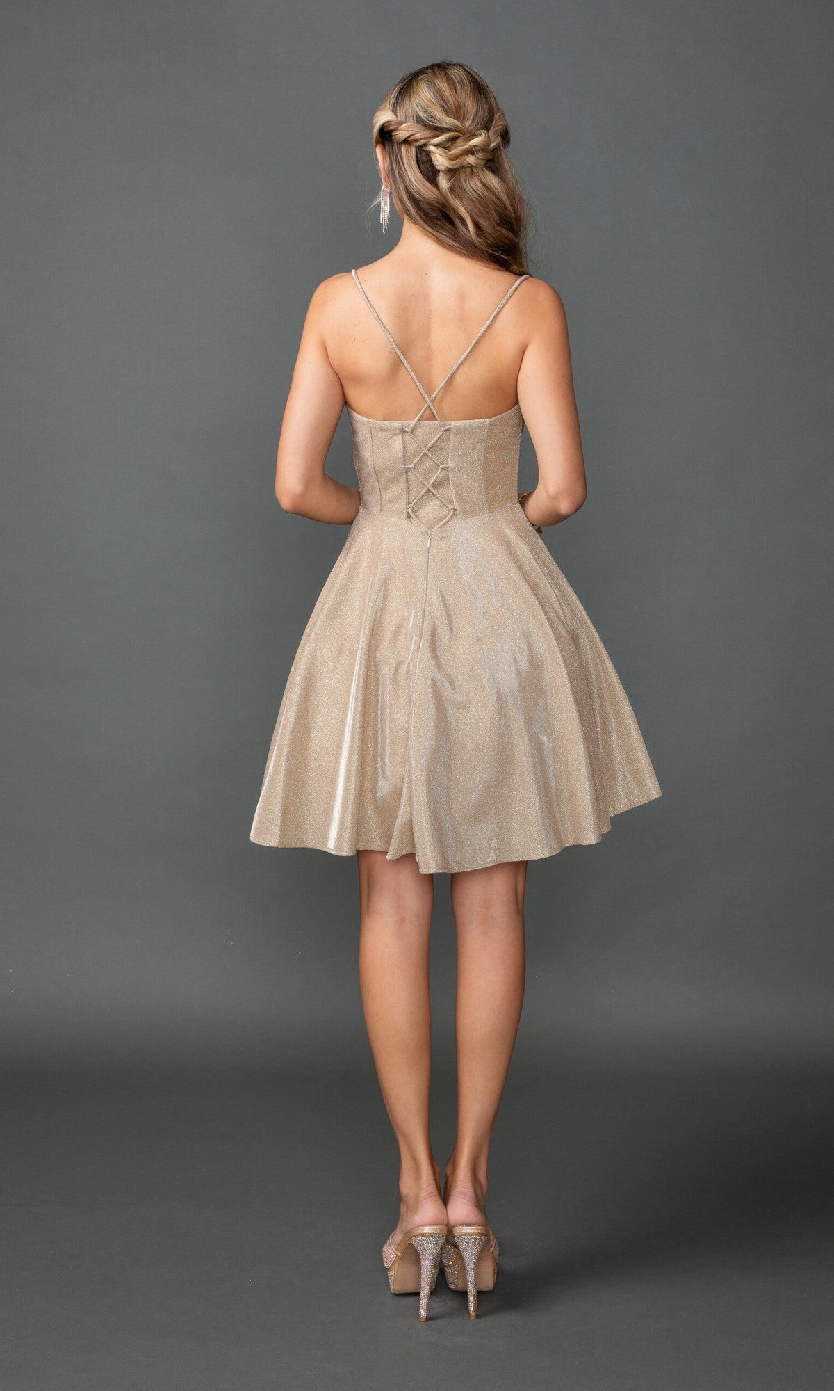 A woman faces away in the Dancing Queen 3421 short prom dress, featuring glitter fabric, crisscross straps, a lace-up back, and corset bodice detail with high heels, set against a plain gray background.